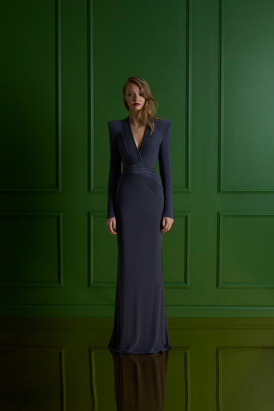 Woman in a long dark blue dress standing against a green paneled wall.